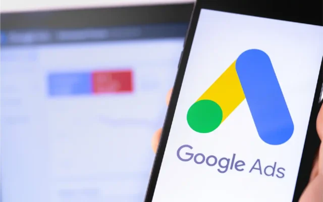 A smartphone displaying the Google Ads logo, with a blurred Google Ads dashboard in the background.